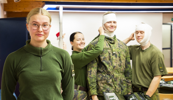 Naissotilas katsoo kameraan, taustalla kolme lääkintälinjan oppilaskorpraalia harjoittelee pään sitomista hymyssä suin.