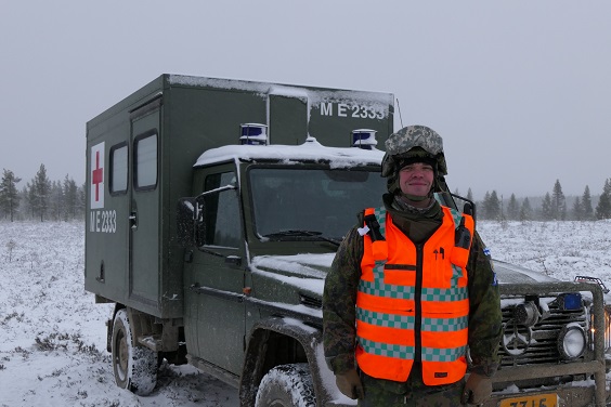 Kuva jossa kenttäsairaanhoitaja maastovarusteissa kypärä päässä maastoambulanssin edessä talvisessa maisemassa.