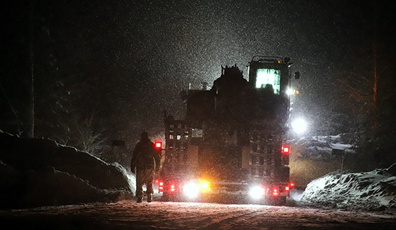 Kaivinkone lavetin päällä ja sotilaan hahmo takaapäin kuvattuna pimeällä lumisateessa maasto-olosuhteissa. Ajoneuvon valot valaisevat pimeää metsää.