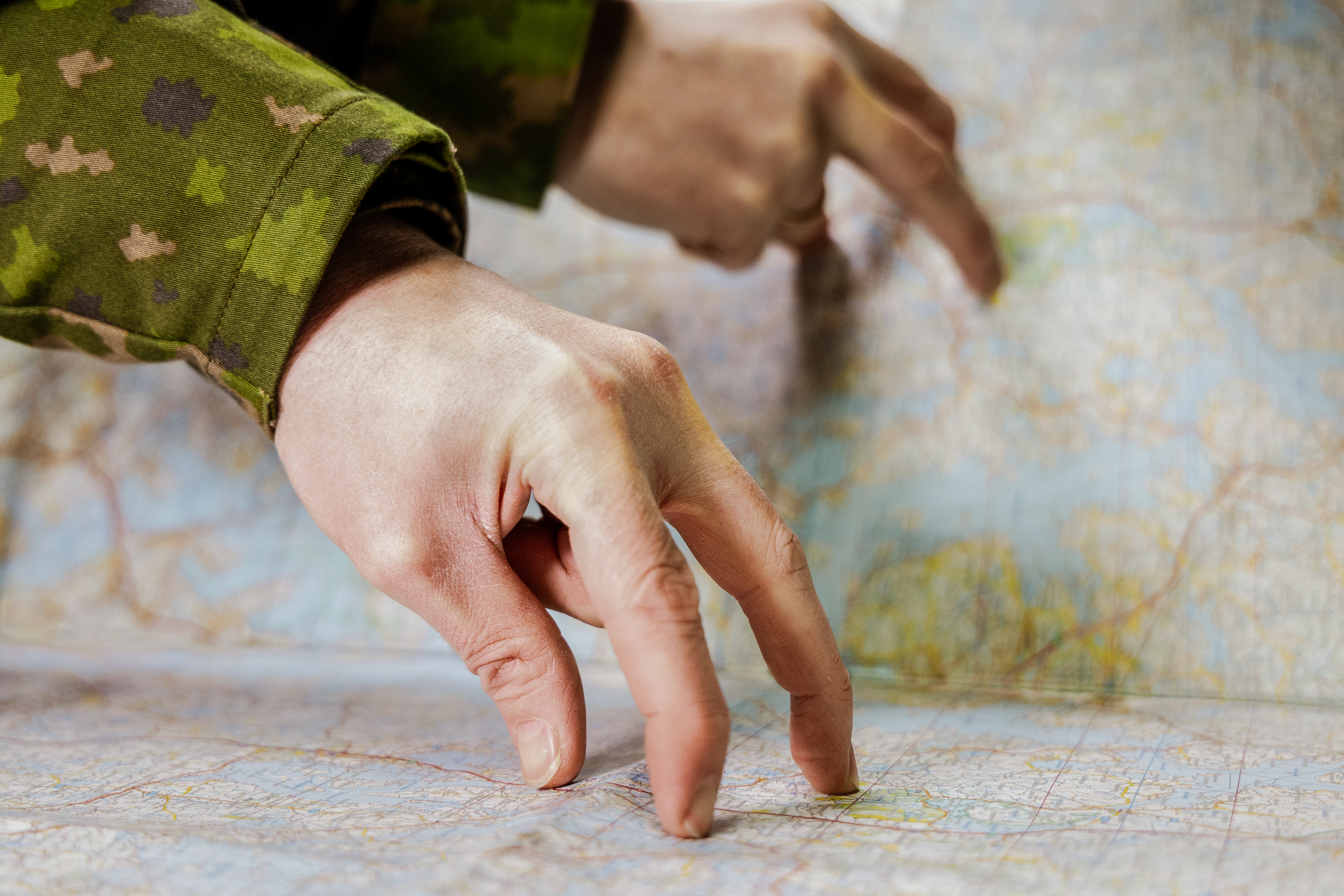 Kuvassa sotilaan käsi kartalla mittaamassa välimatkoja. Sotilaspuvun hiha näkyy, kartta on alapuolella ja ympärillä.