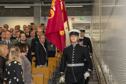 Ihmiset nousevat seisomaan salissa, kun Maanpuolustuskorkeakoulun lippu kannetaan sisään.