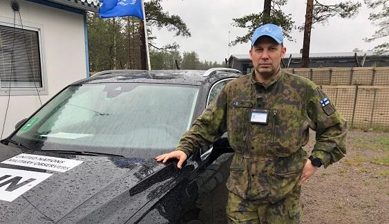 Maastopuvussa päällä oleva Peltola seisoo auton vieressä YK:n sininen lippalakki päässä