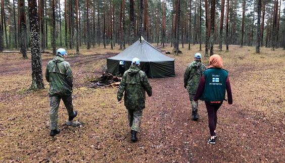 Kolme maastopukuista sotilasta ja siviili kävelevät metsässä kohti telttaa